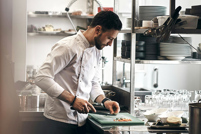 Cocinero profesional con uniforme blanco cortando verduras en una cocina industrial impecable, representando los estándares de higiene y seguridad alimentaria enseñados en el curso de manipulador de alimentos de Cosaplag.